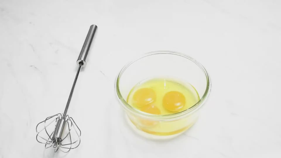 Semi-automatic stainless steel egg beater whisk spinning rapidly while mixing eggs in a bowl.